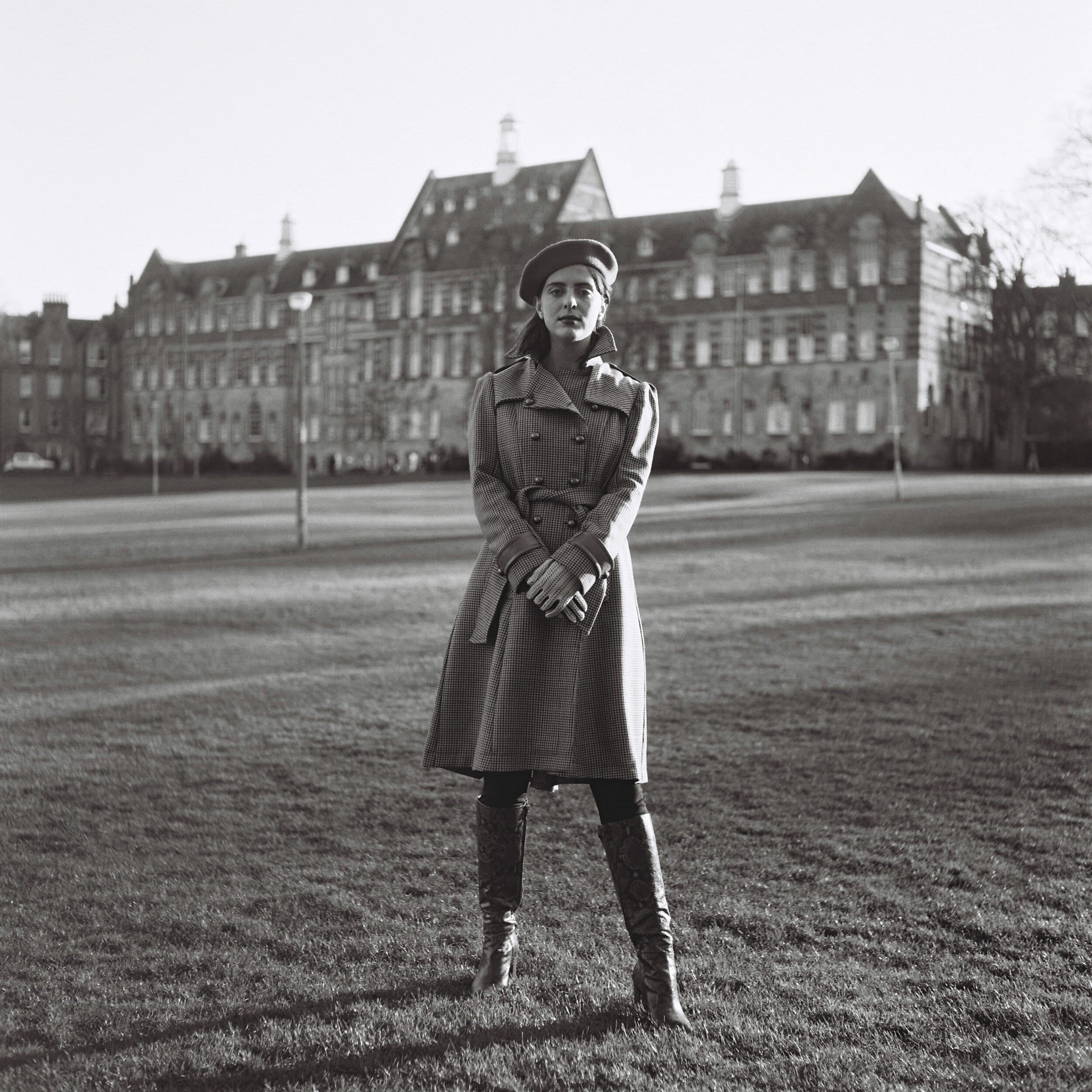Person in a coat and hat standing on grass with a building in the background
