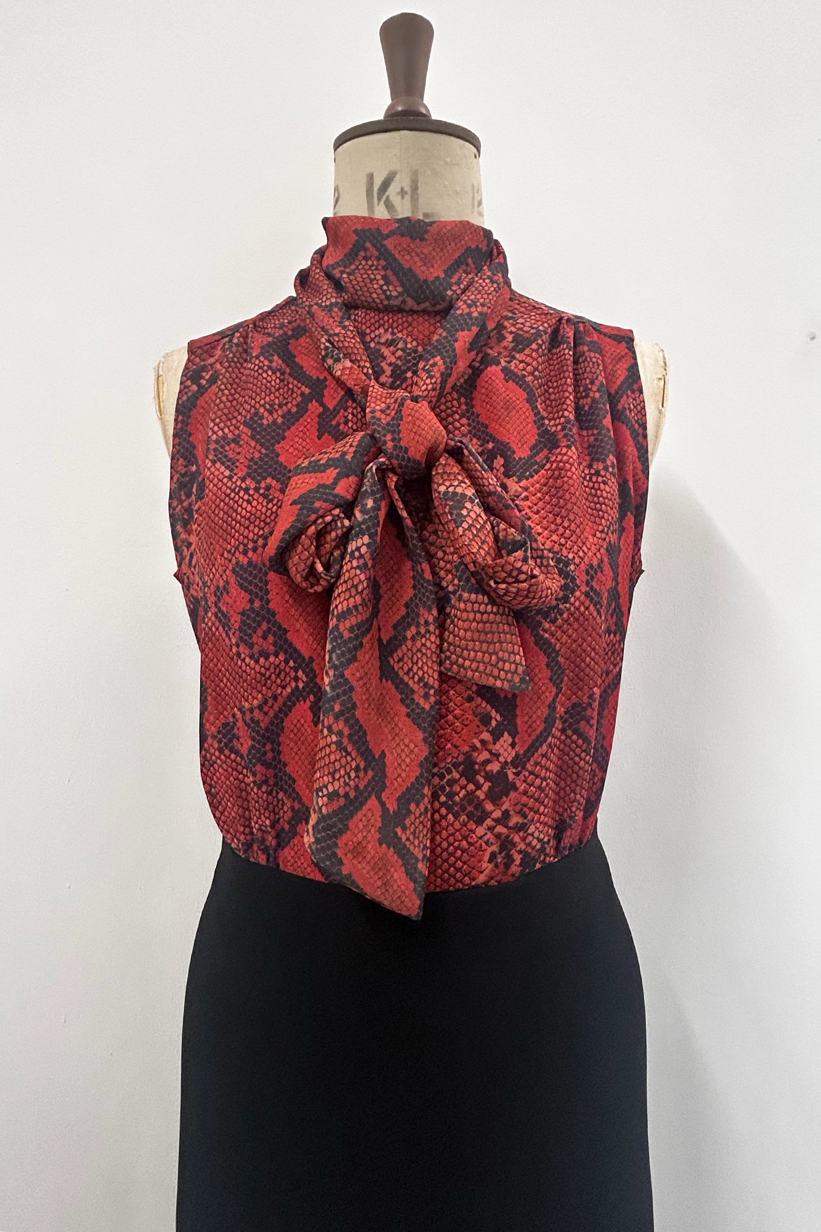A sleeveless high-neck top with a large front bow, featuring a red and black snakeskin pattern, displayed on a dress form mannequin against a plain white wall.