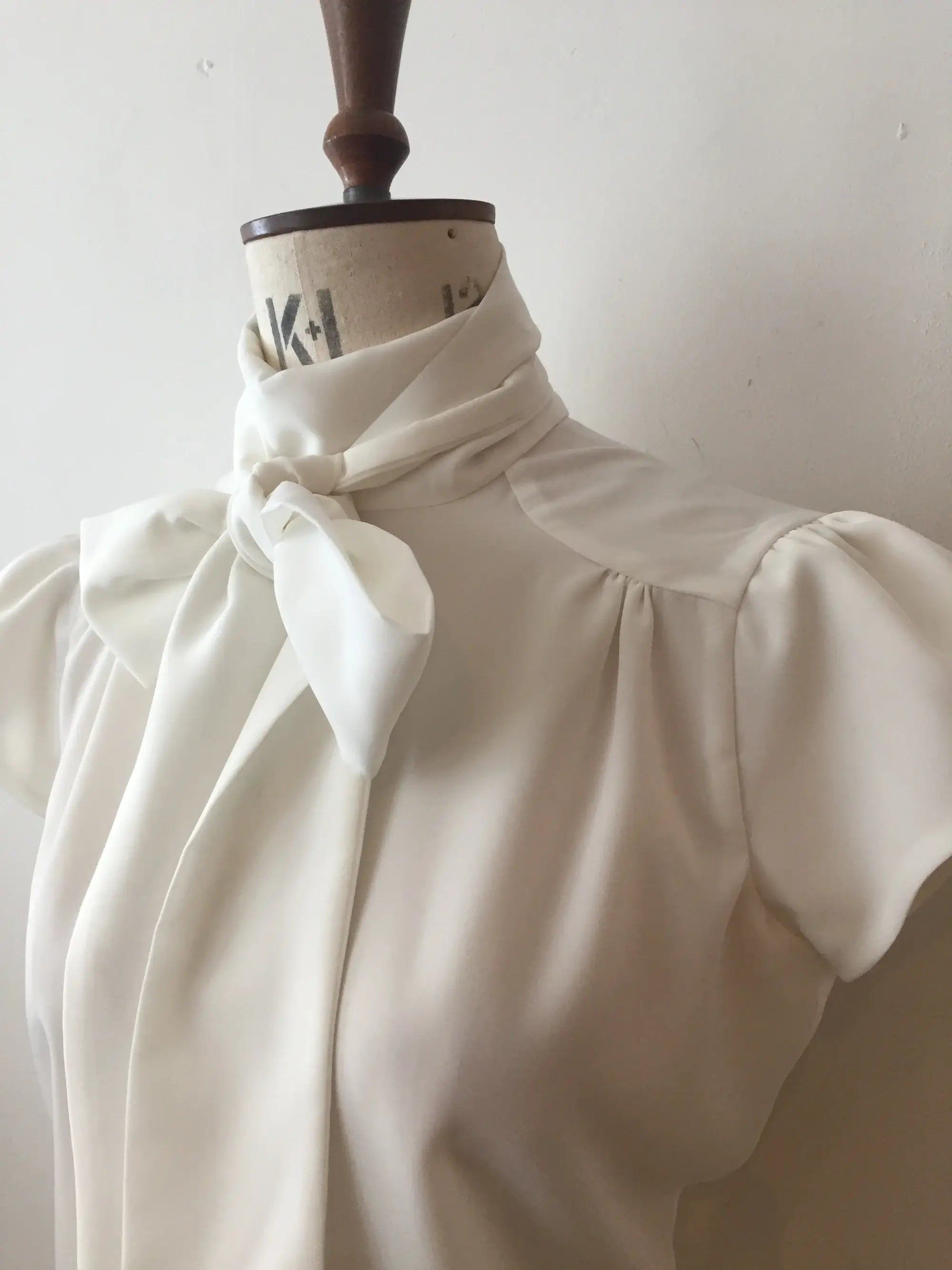 A white blouse with cap sleeves and a silky tie neck displayed on a mannequin.