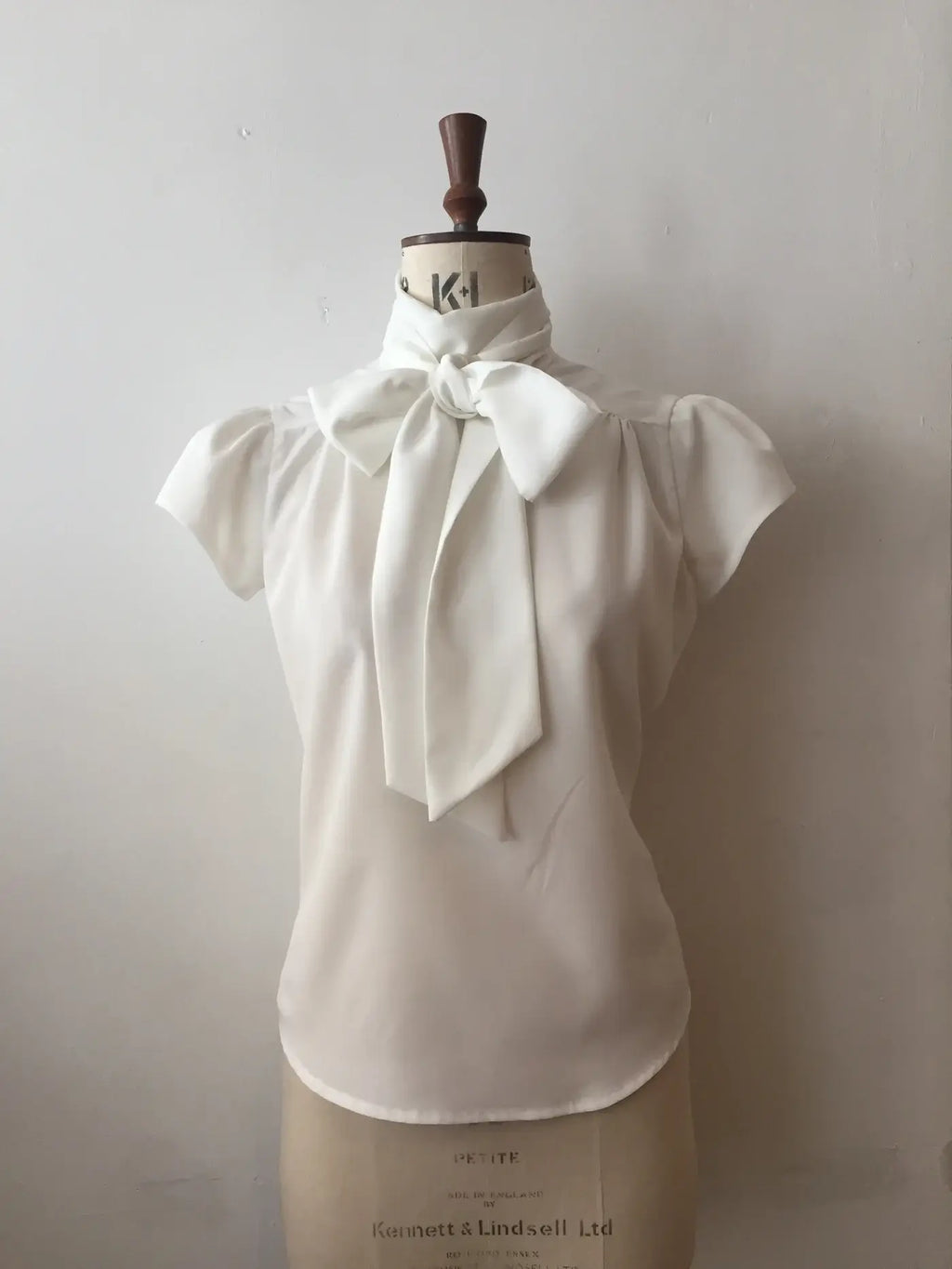 A white blouse with cap sleeves and a silky tie-neck displayed on a mannequin.