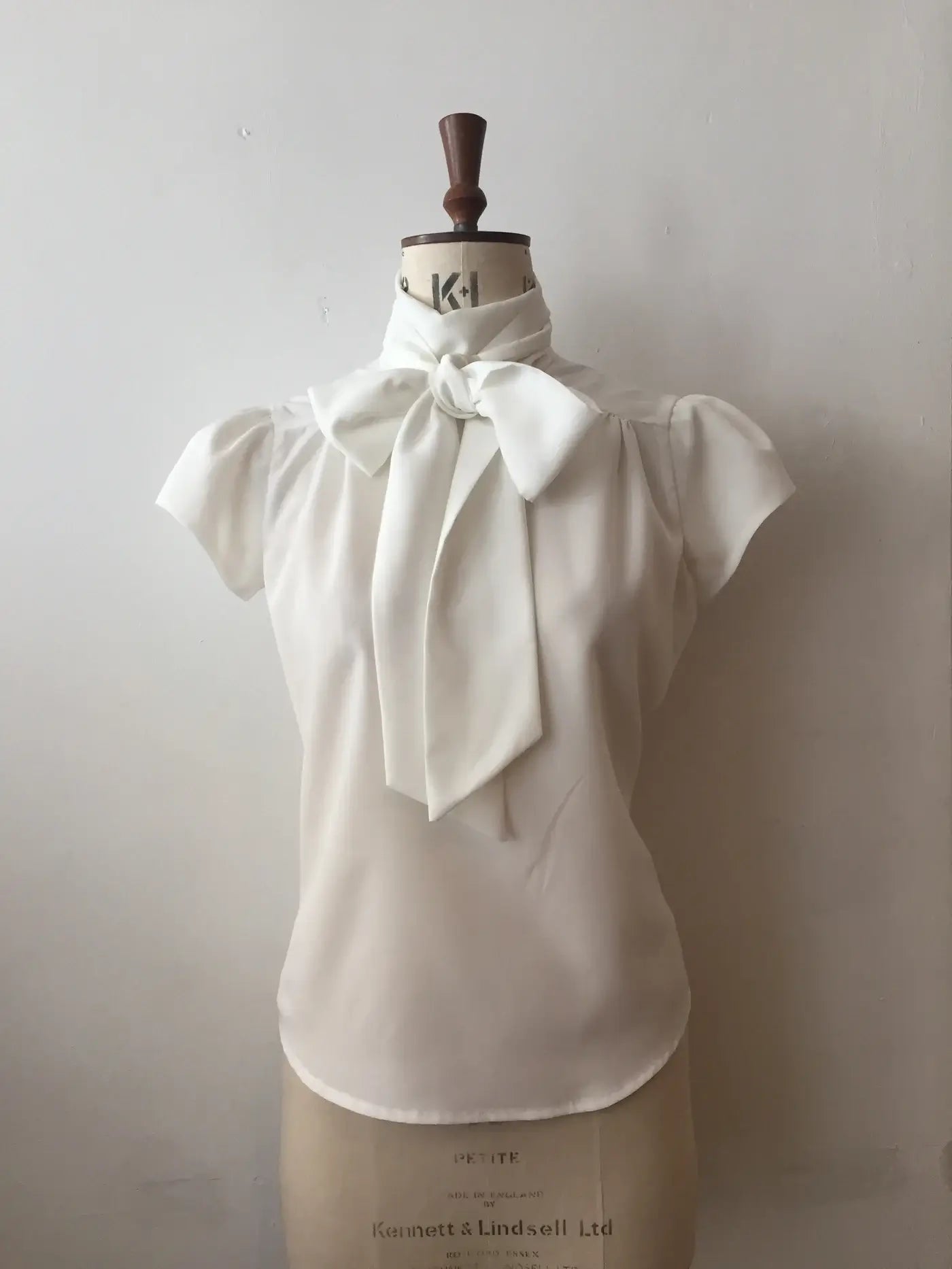 A white blouse with cap sleeves and a silky tie-neck displayed on a mannequin.