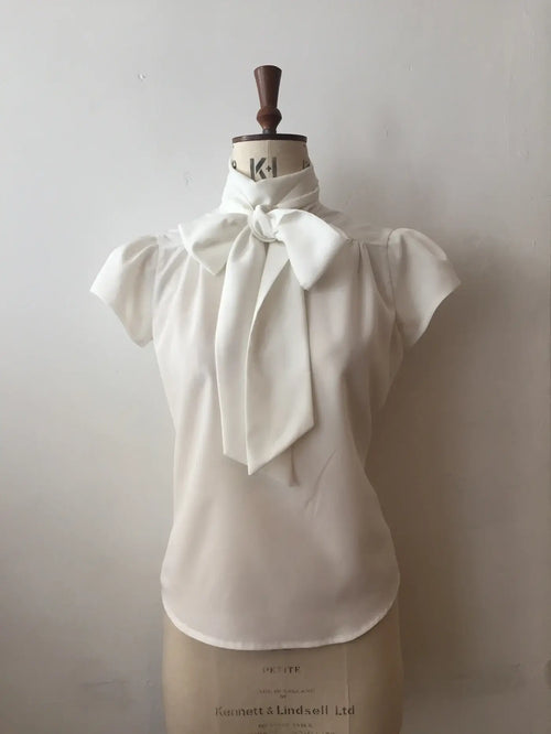 A white blouse with cap sleeves and a silky tie-neck displayed on a mannequin.