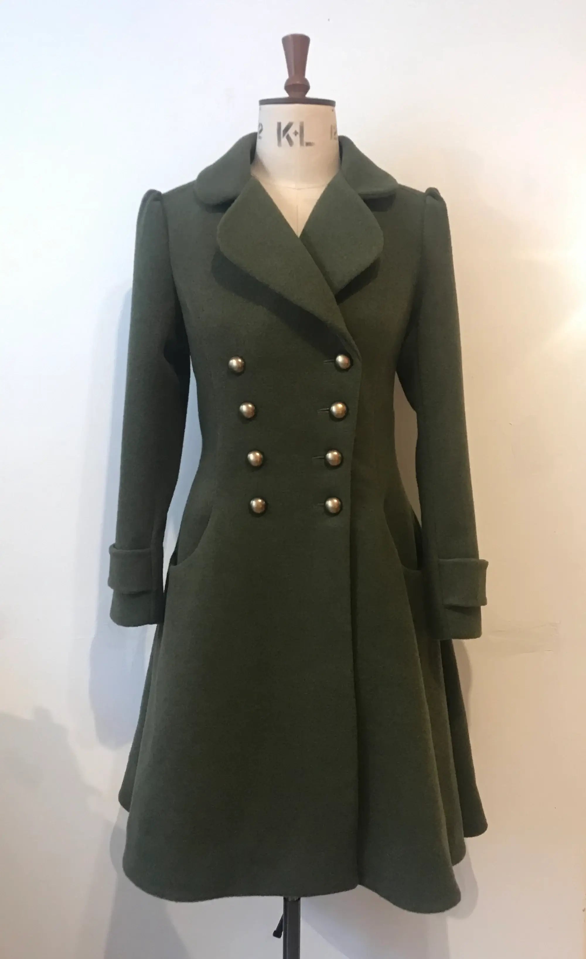 A double-breasted, flared green coat with antique brass-effect buttons and a soft, rounded collar, displayed on a mannequin.