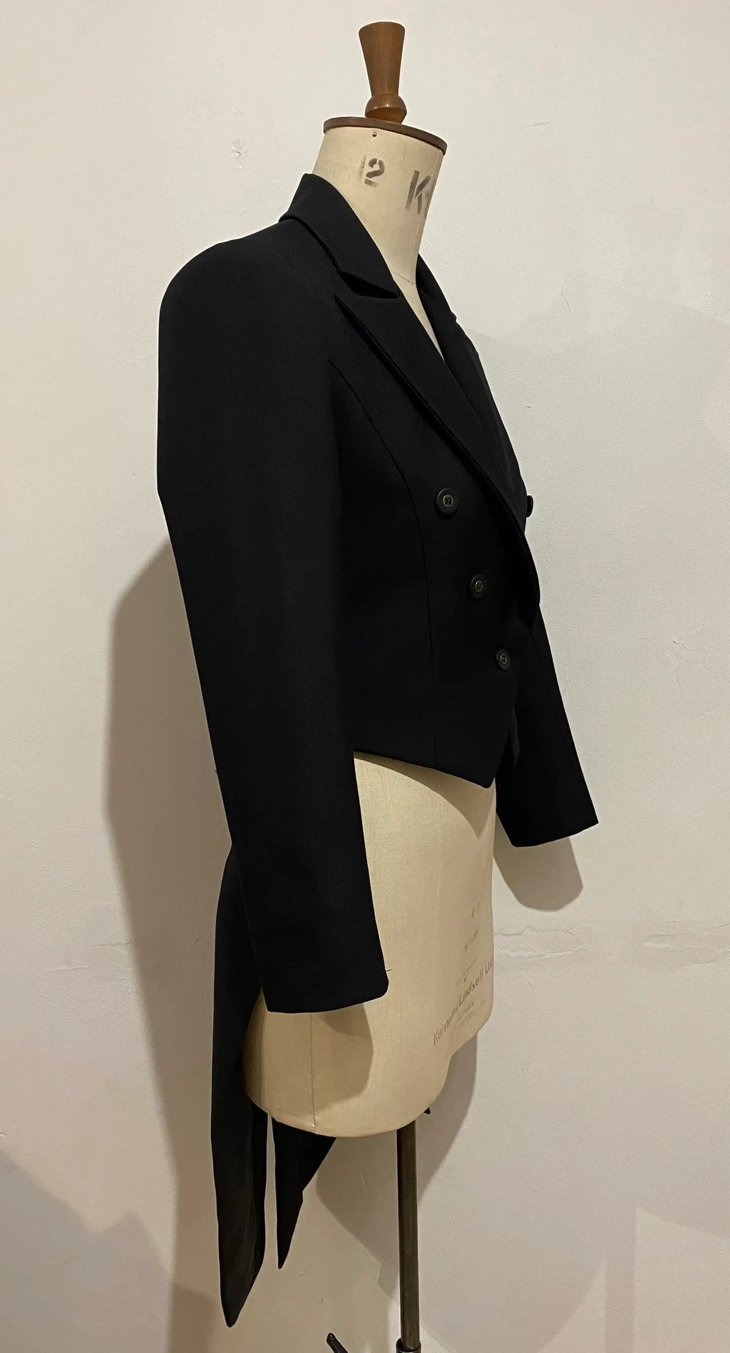 A black tailcoat displayed on a mannequin, featuring a traditional design with strong shoulders and a dramatic silhouette.