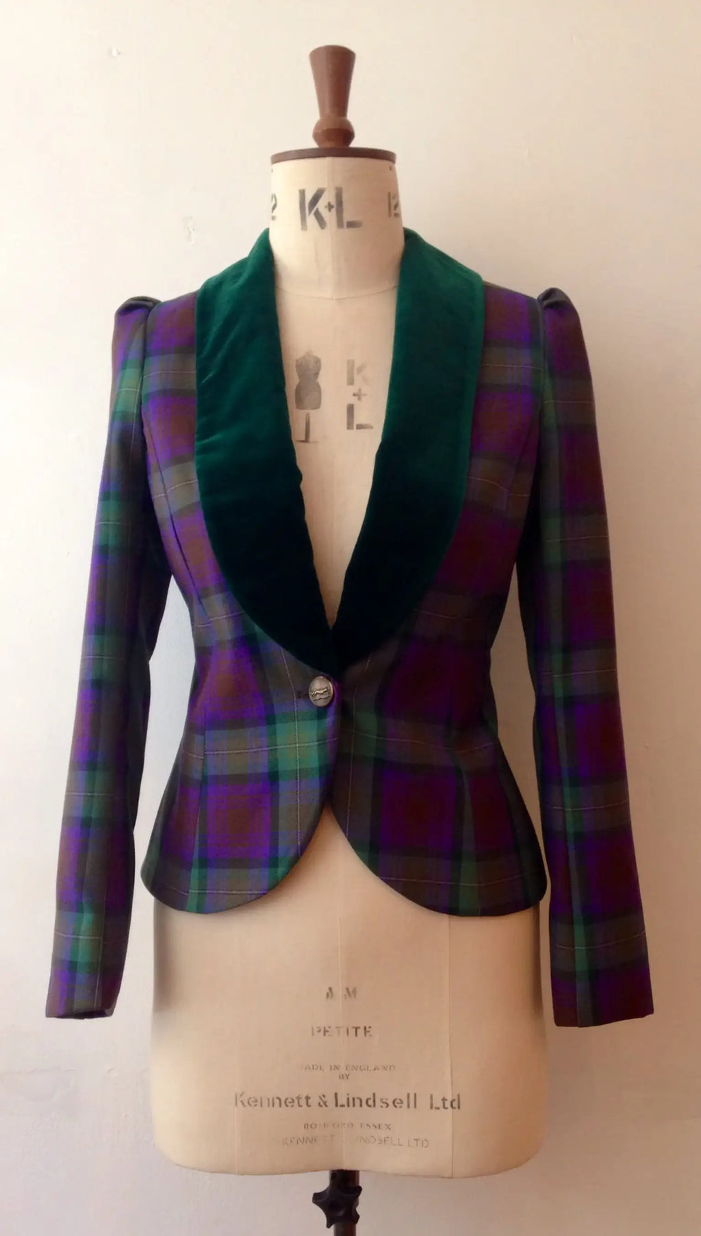 A velvet and tartan show jacket displayed on a mannequin, with the mannequin marked with 'KL' initials. The jacket has a classic silhouette with a green velvet collar and trim, and a multicolored tartan pattern.